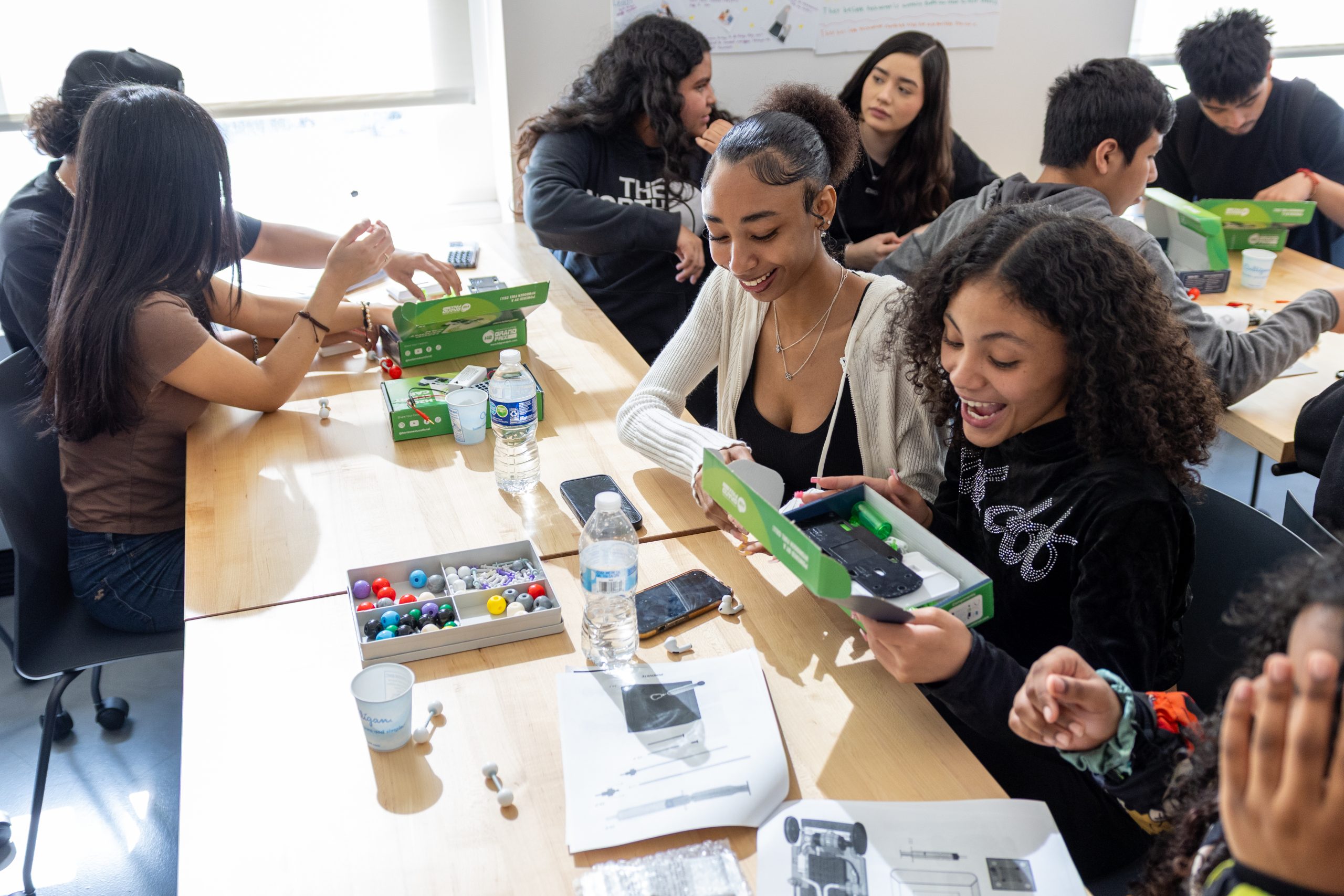 Students from Western International High School sit around bright wooden tables, actively engaged in a workshop led by the University of Michigan Hydrogen Race Team. In the foreground, two smiling teenage girls open a green box containing a hydrogen-propelled toy car kit, looking excited and happy. The table is scattered with instruction sheets, water bottles, and trays of colorful molecule models.