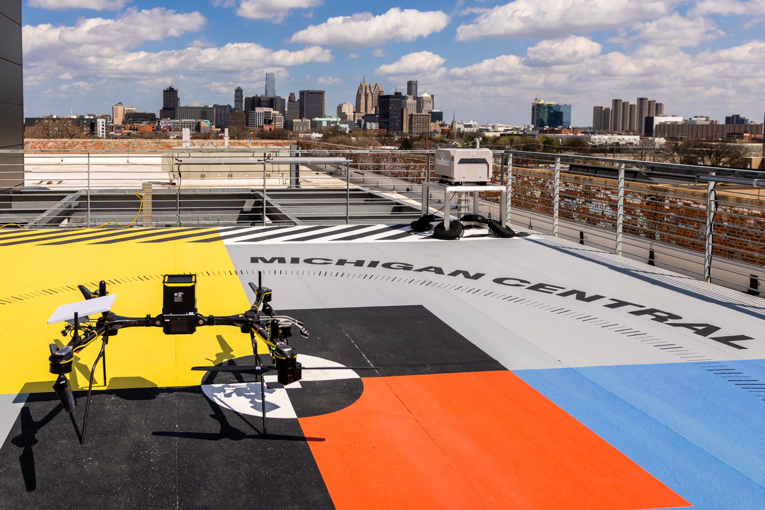 A multi-rotor drone sits on the colorful rooftop landing pad of Michigan Central in Detroit, with the city's downtown skyline visible in the background.